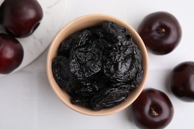 Delicious prunes in bowl and fresh ripe plums on white table, flat lay