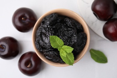 Photo of Delicious prunes with mint in bowl and fresh ripe plums on white table, flat lay