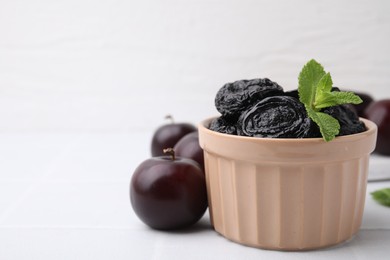 Photo of Delicious prunes with mint in bowl and fresh ripe plums on white table, closeup. Space for text