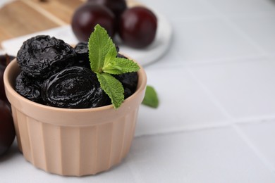 Photo of Delicious prunes with mint in bowl and fresh ripe plums on white tiled table, closeup. Space for text