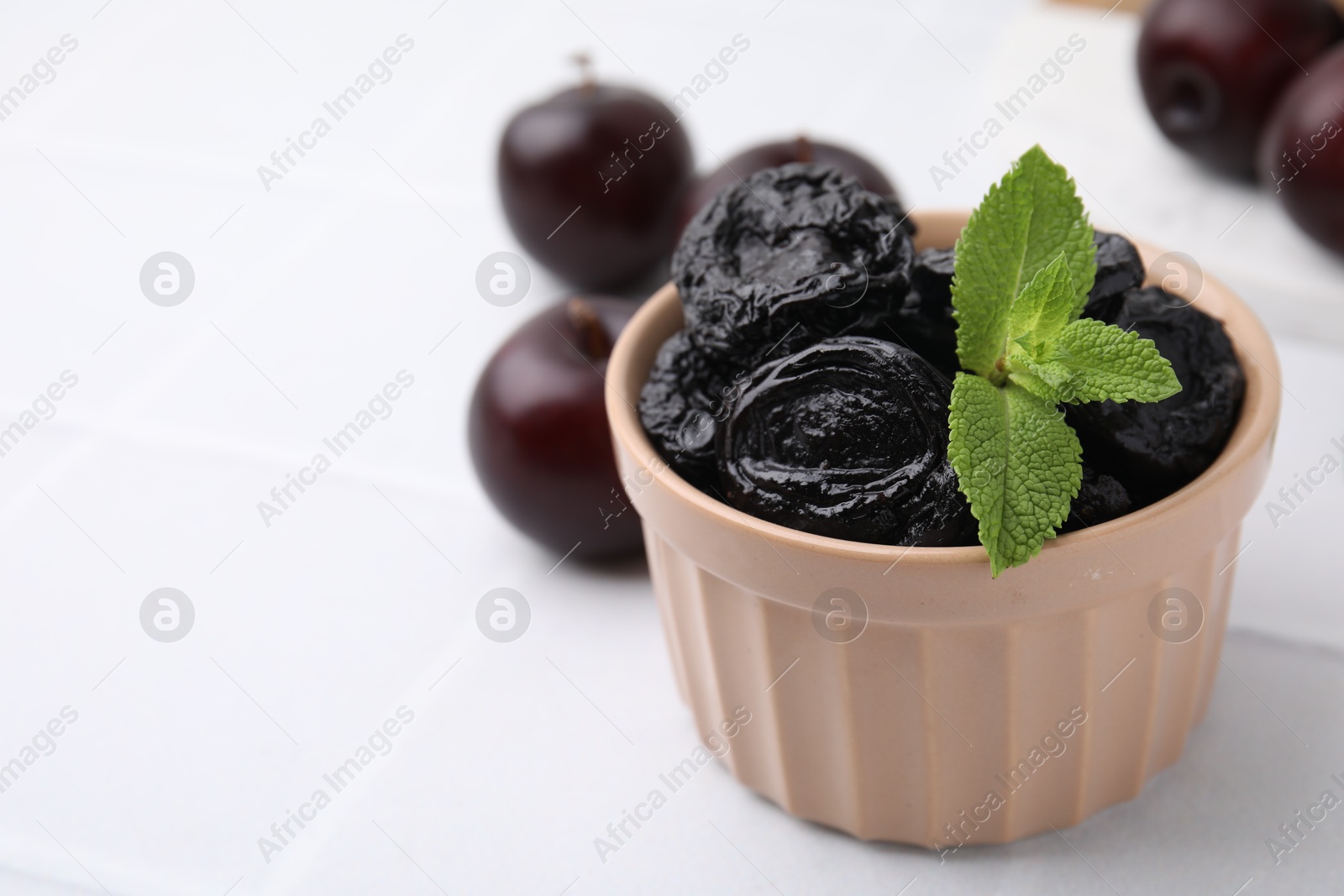 Photo of Delicious prunes with mint in bowl and fresh ripe plums on white table, closeup. Space for text