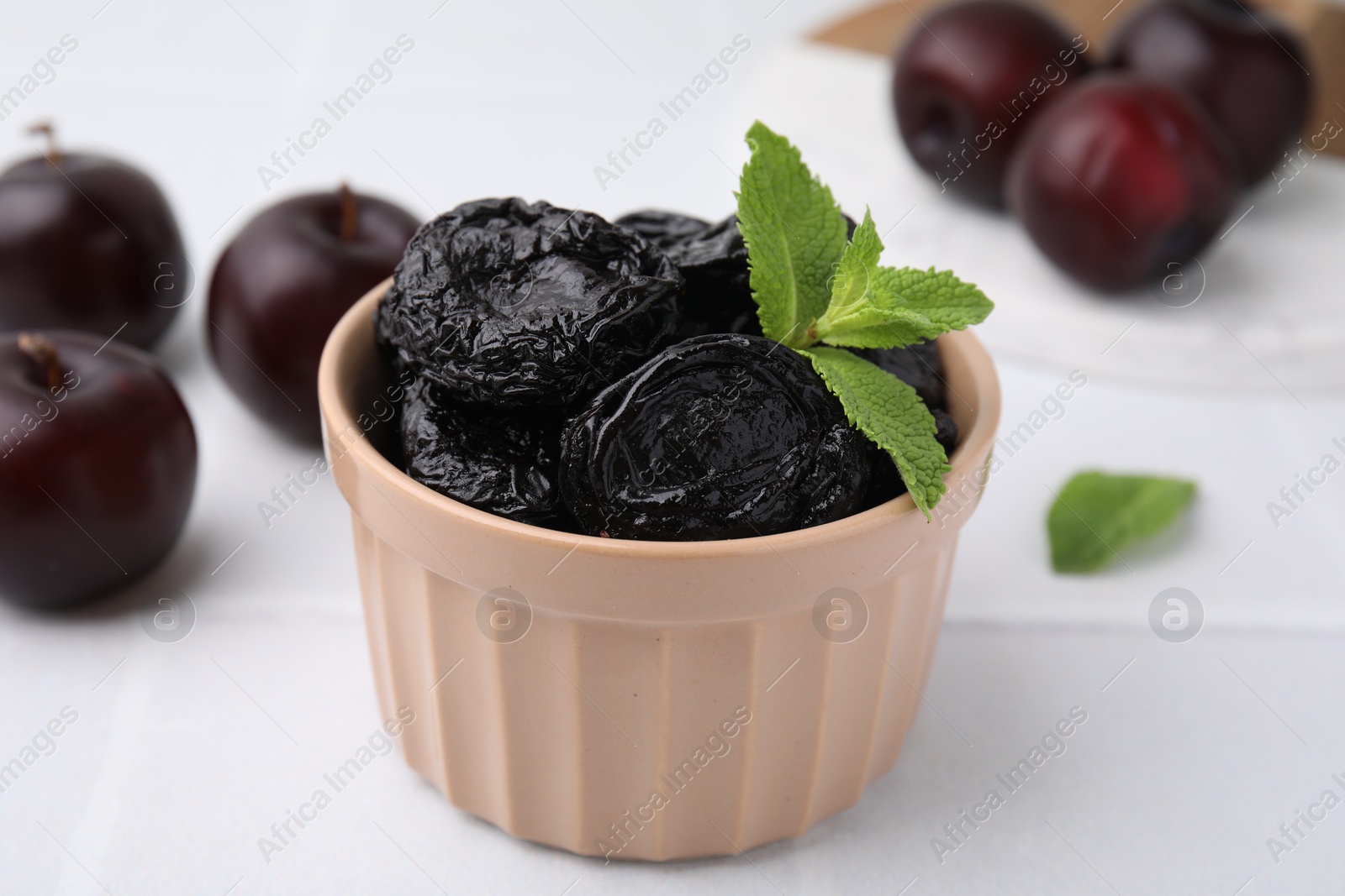 Photo of Delicious prunes with mint in bowl and fresh ripe plums on white table, closeup