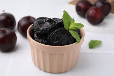 Delicious prunes with mint in bowl and fresh ripe plums on white table, closeup