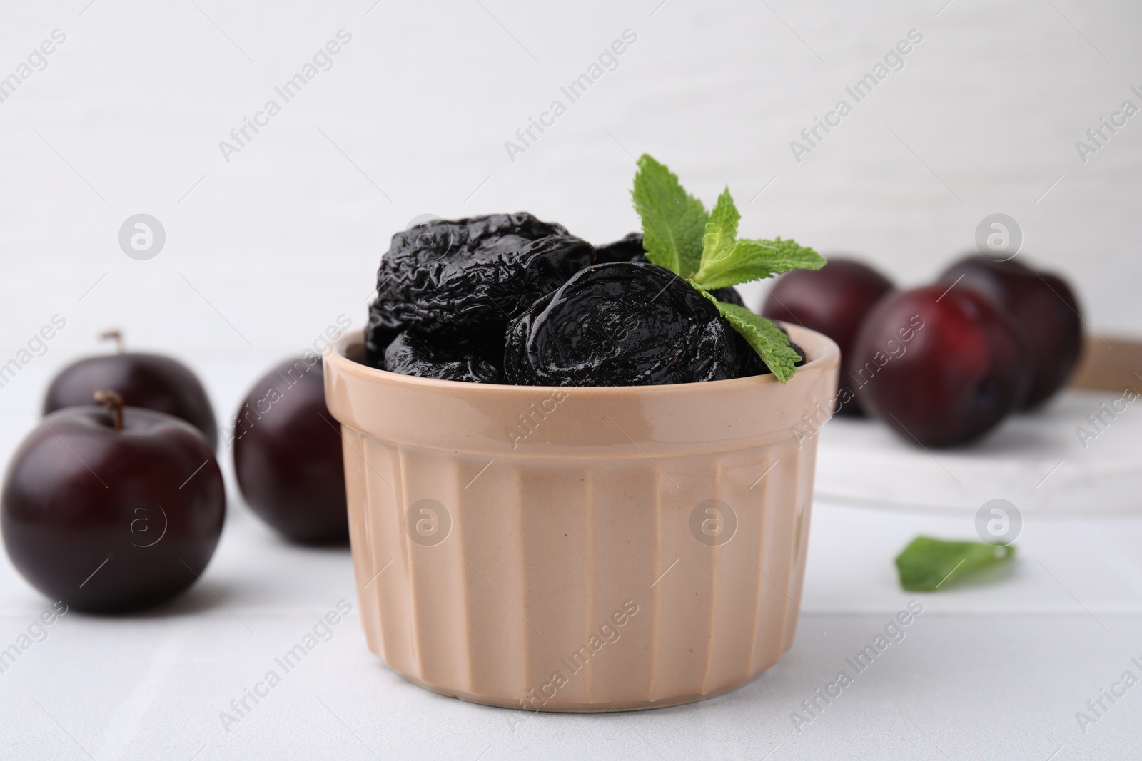 Photo of Delicious prunes with mint in bowl and fresh ripe plums on white table, closeup