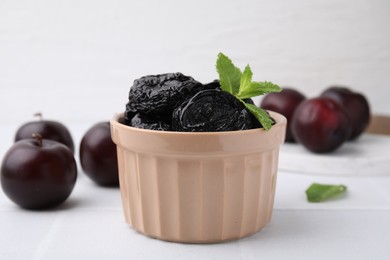 Delicious prunes with mint in bowl and fresh ripe plums on white table, closeup