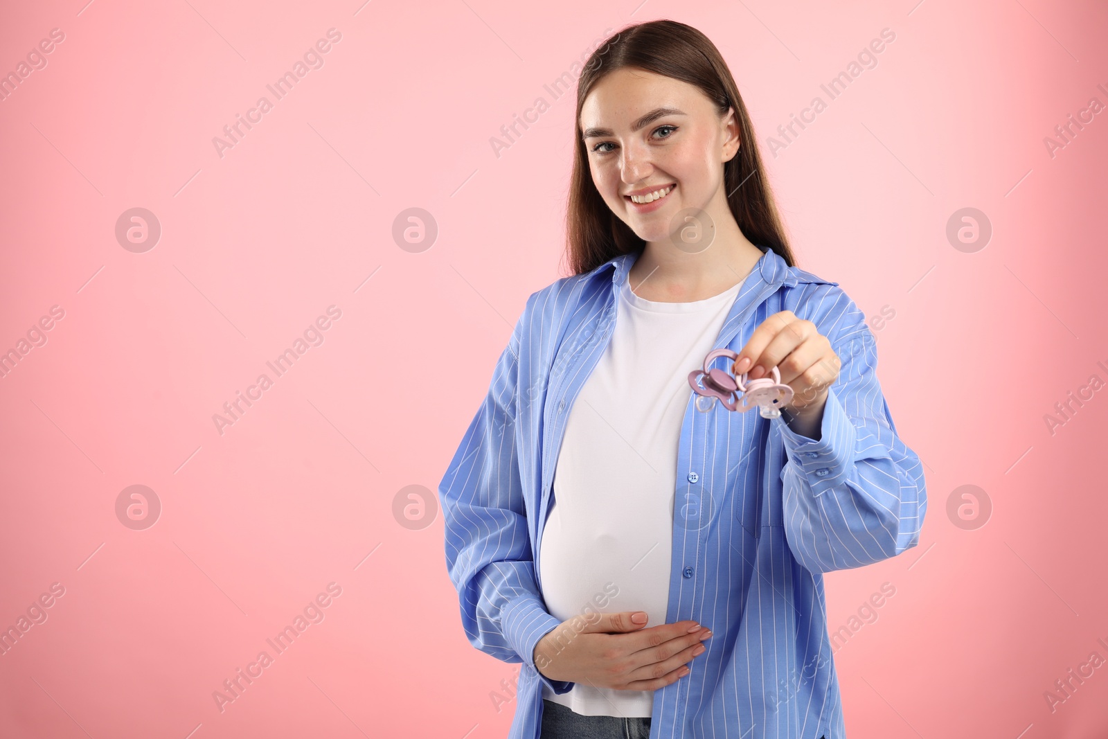 Photo of Expecting twins. Pregnant woman holding two pacifiers on pink background, space for text