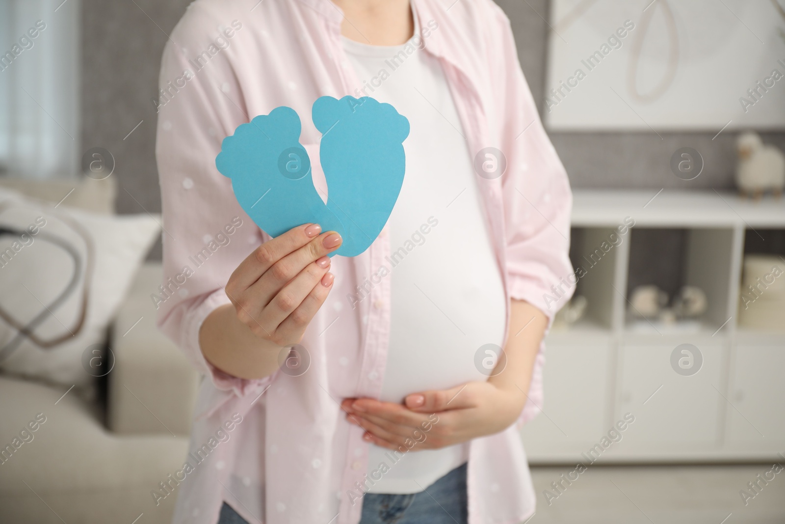 Photo of Expecting twins. Pregnant woman holding two paper cutouts of feet at home, closeup