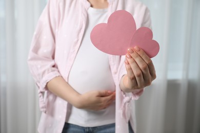 Photo of Expecting twins. Pregnant woman holding two paper cutouts of hearts at home, closeup