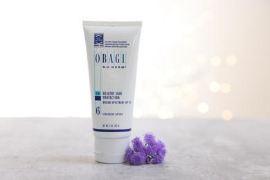 Photo of Leiden, Netherlands - June 11, 2024: Tube of Obagi sunscreen and ageratum flower on gray table against light background with blurred lights