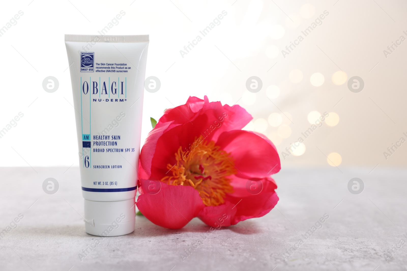 Photo of Leiden, Netherlands - June 11, 2024: Tube of Obagi sunscreen and peony flower on gray table against light background with blurred lights