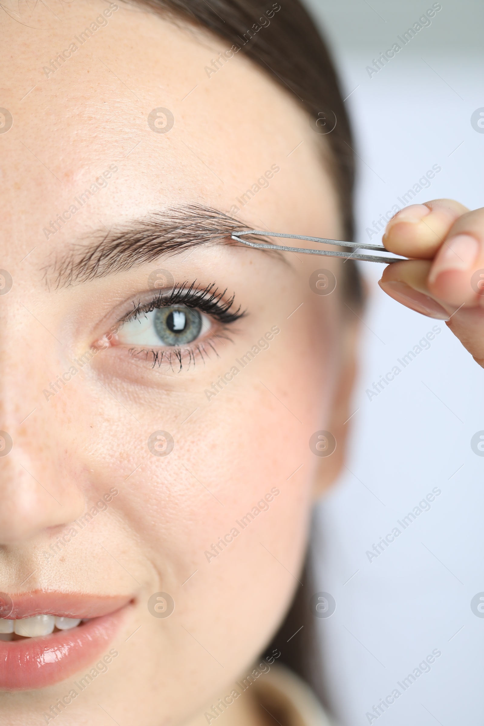 Photo of Young woman plucking eyebrow with tweezers on light background, closeup