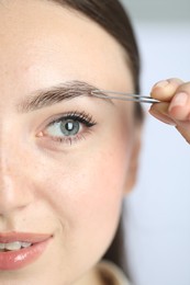 Young woman plucking eyebrow with tweezers on light background, closeup