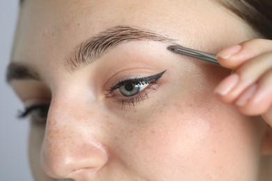 Young woman plucking eyebrow with tweezers on light background, closeup