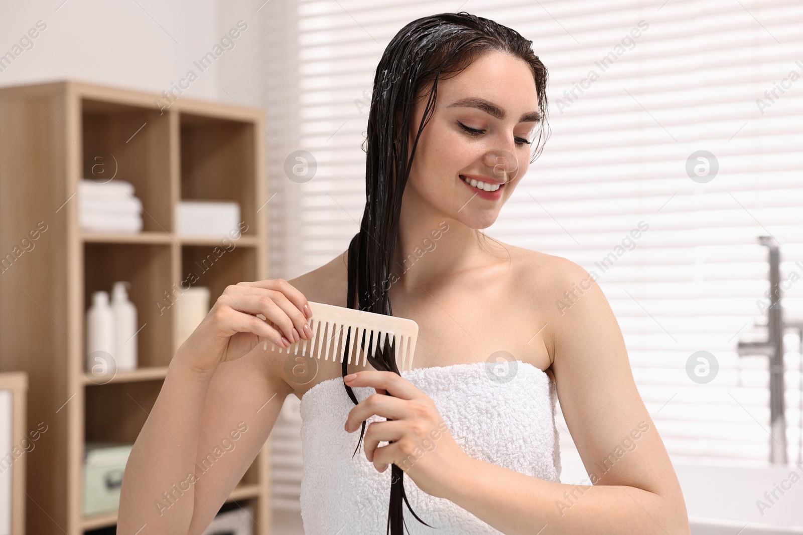 Photo of Smiling woman combing her hair with applied mask in bathroom