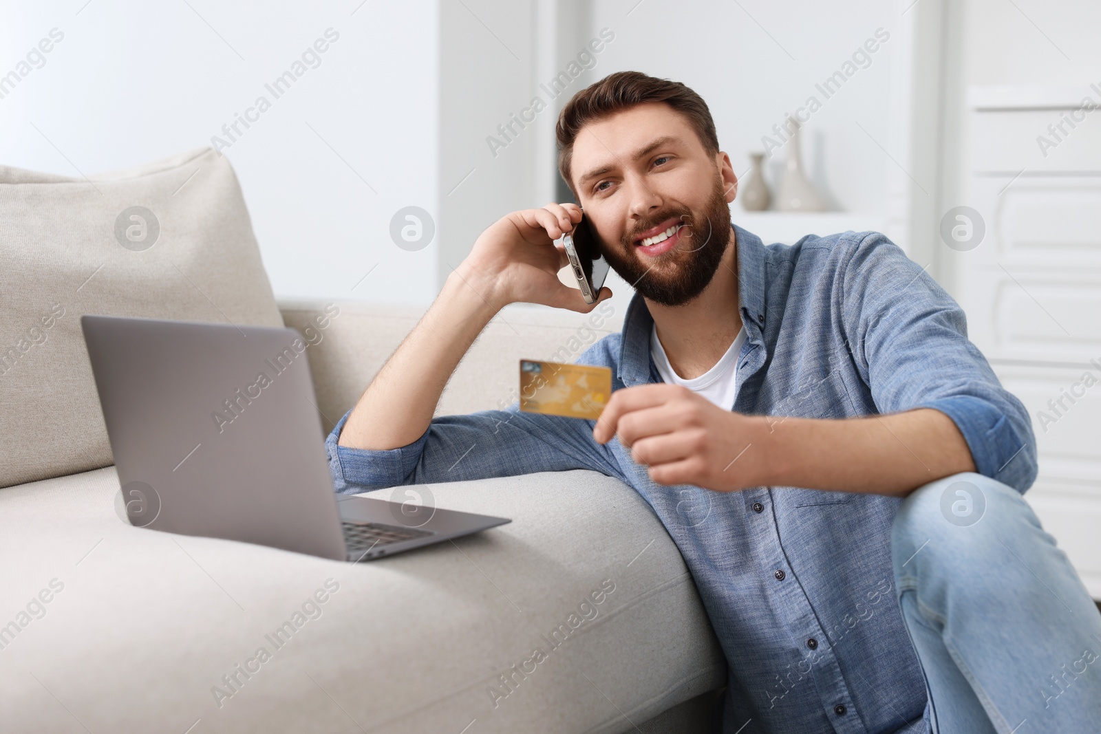 Photo of Online banking. Happy young man with credit card talking by smartphone at home