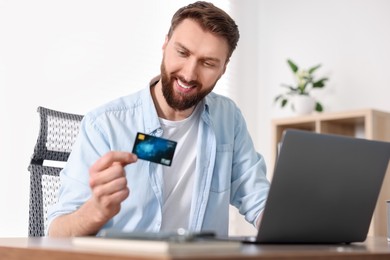 Photo of Online banking. Happy young man with credit card and laptop paying purchase at table indoors