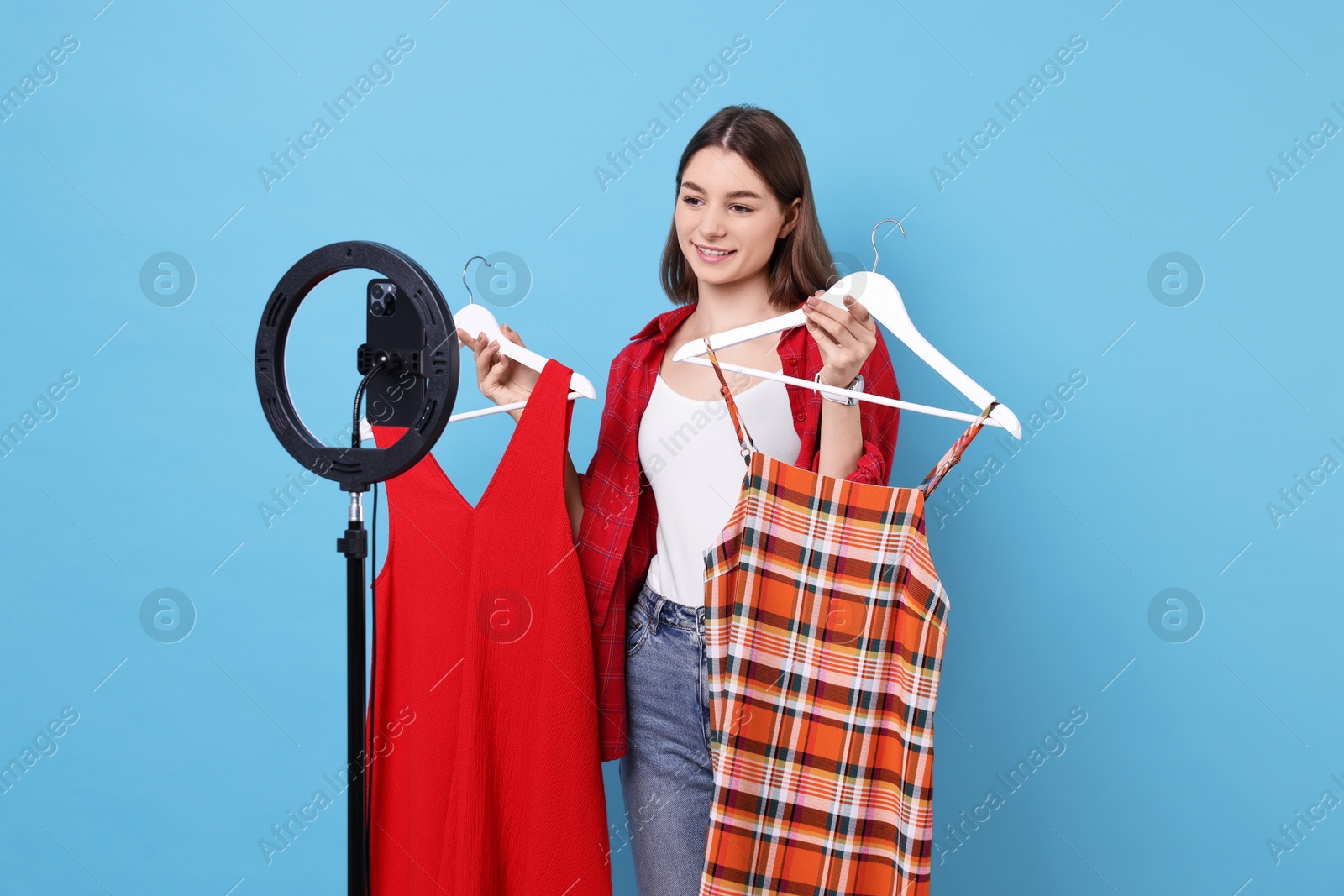 Photo of Fashion blogger reviewing dresses and recording video with smartphone and ring lamp on light blue background