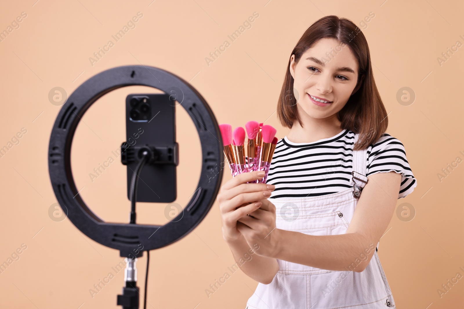 Photo of Beauty blogger reviewing makeup brushes and recording video with smartphone and ring lamp on beige background