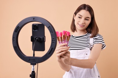 Beauty blogger reviewing makeup brushes and recording video with smartphone and ring lamp on beige background