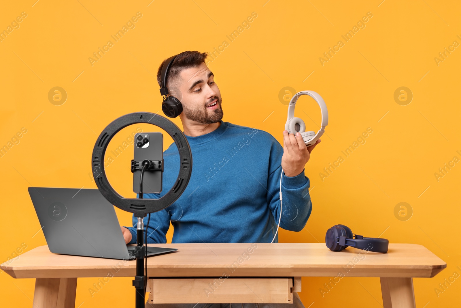 Photo of Technology blogger reviewing headphones and recording video with smartphone and ring lamp at wooden table on orange background