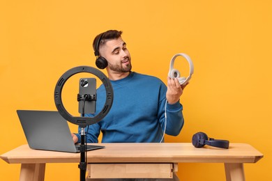 Technology blogger reviewing headphones and recording video with smartphone and ring lamp at wooden table on orange background