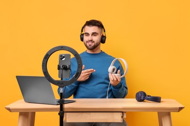 Technology blogger reviewing headphones and recording video with smartphone and ring lamp at wooden table on orange background