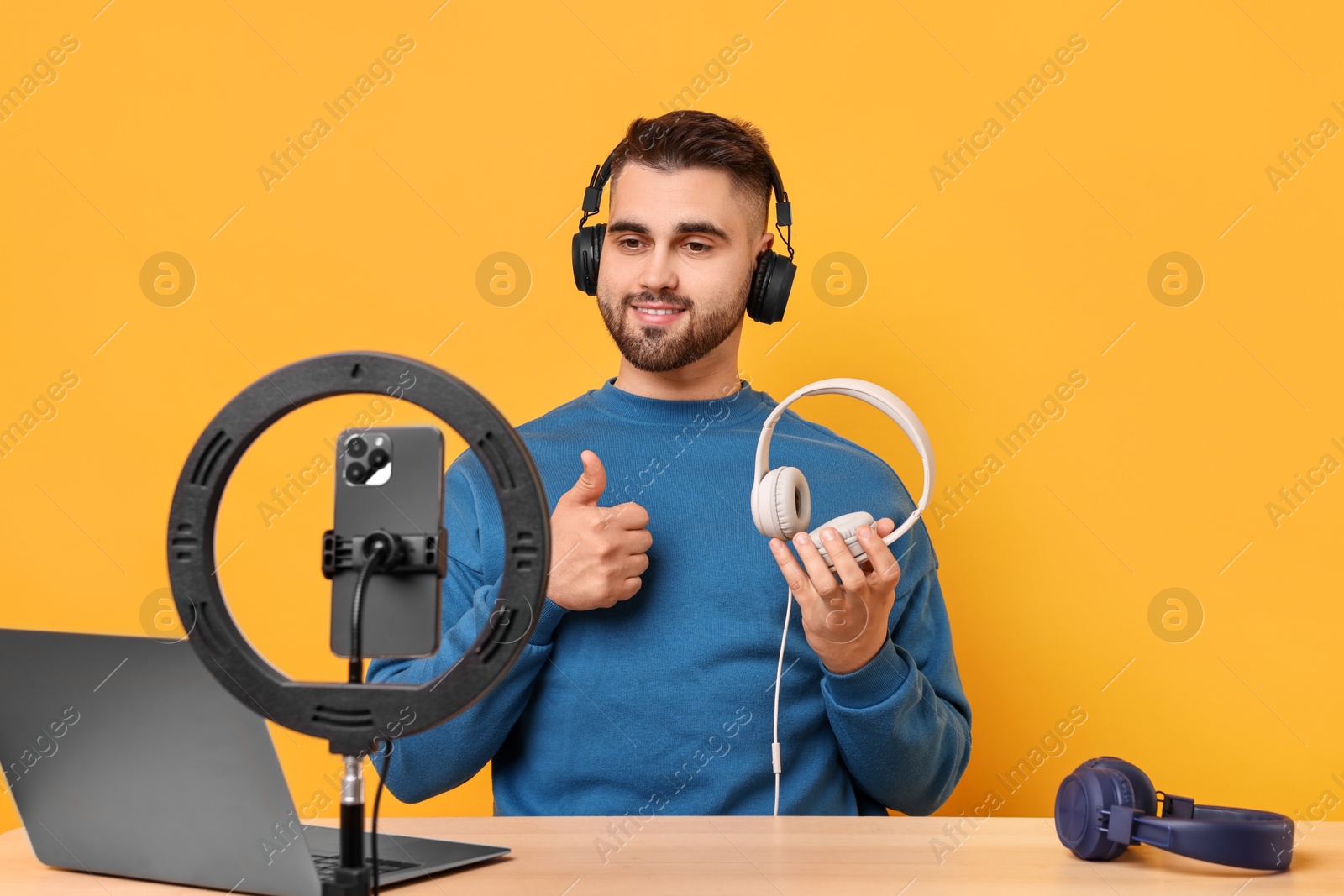 Photo of Technology blogger reviewing headphones and recording video with smartphone and ring lamp at wooden table on orange background