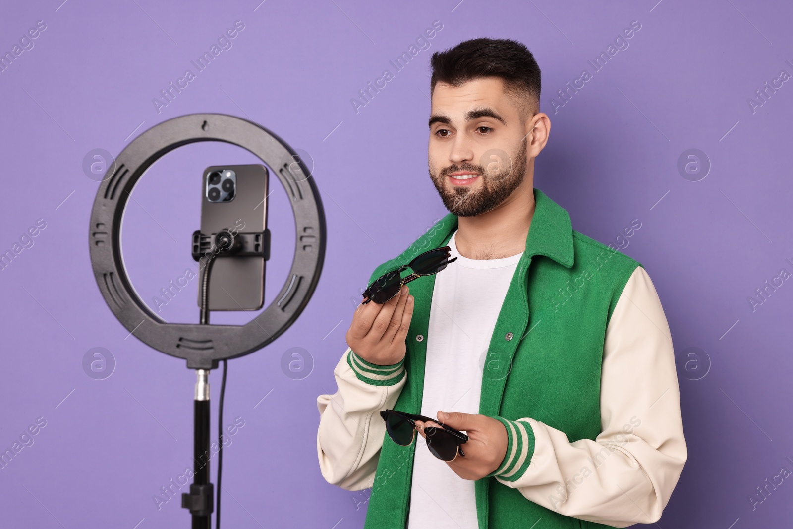 Photo of Fashion blogger reviewing sunglasses and recording video with smartphone and ring lamp on purple background