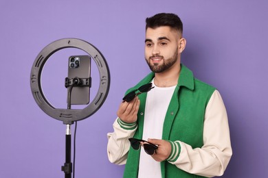 Photo of Fashion blogger reviewing sunglasses and recording video with smartphone and ring lamp on purple background