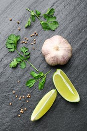 Fresh coriander leaves, dried seeds, garlic and lime wedges on black textured table, flat lay