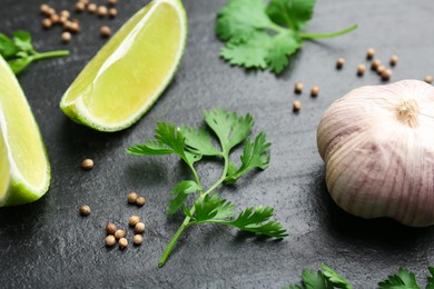 Photo of Fresh coriander leaves, dried seeds, garlic and lime wedges on black textured table, closeup