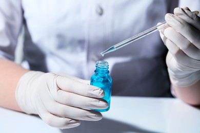 Scientist dripping liquid from pipette into glass bottle at white table, closeup