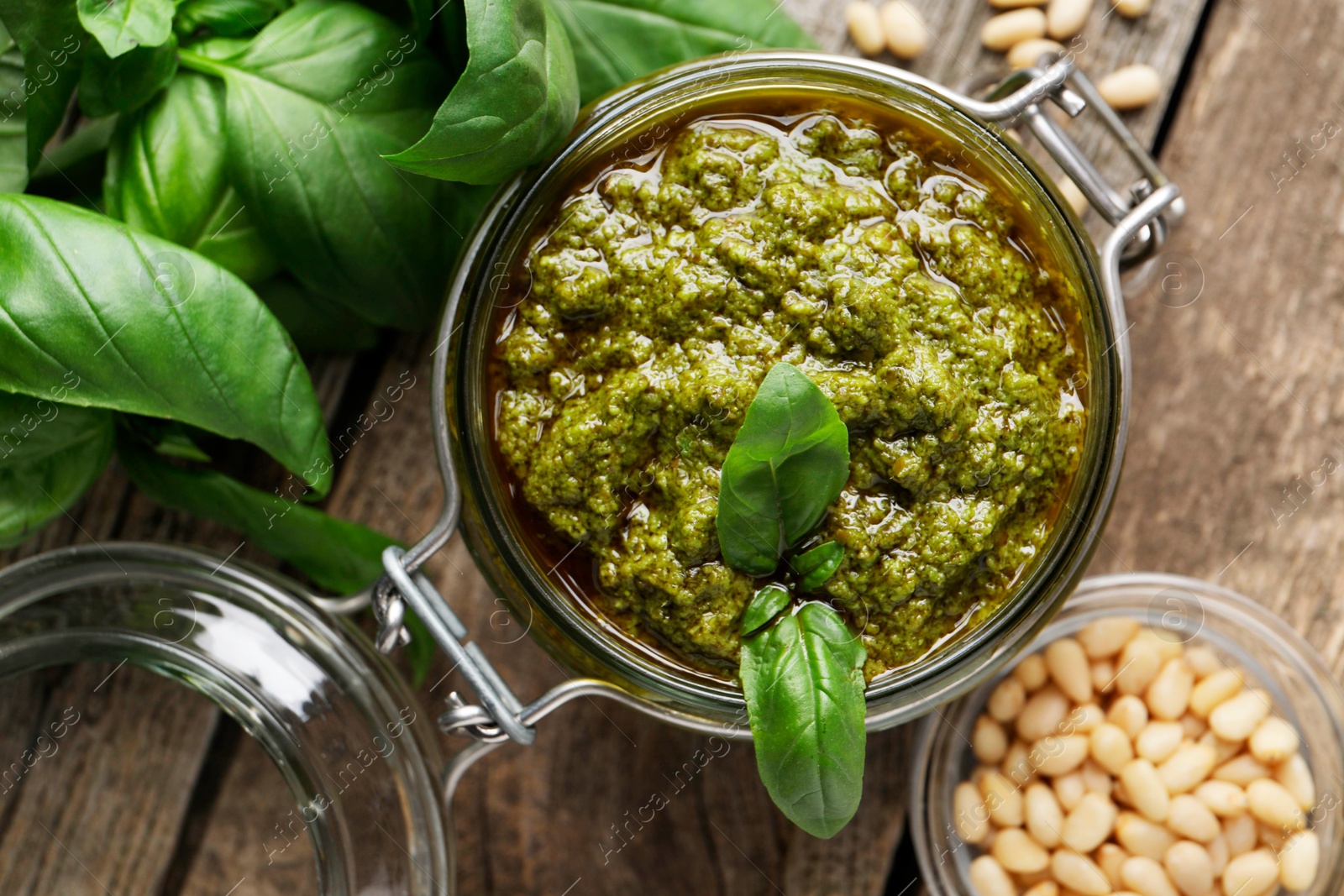 Photo of Tasty pesto sauce in jar, pine nuts and basil on wooden table, top view