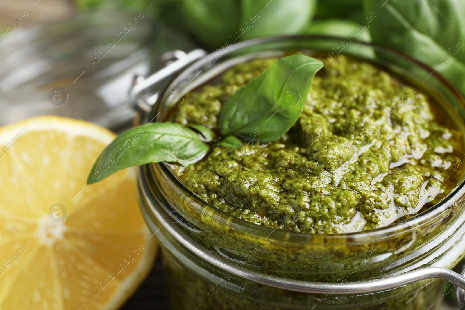 Photo of Tasty pesto sauce in glass jar, basil and lemon on table, closeup