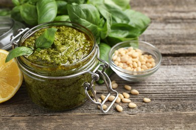 Photo of Tasty pesto sauce in jar, pine nuts, basil and lemon on wooden table