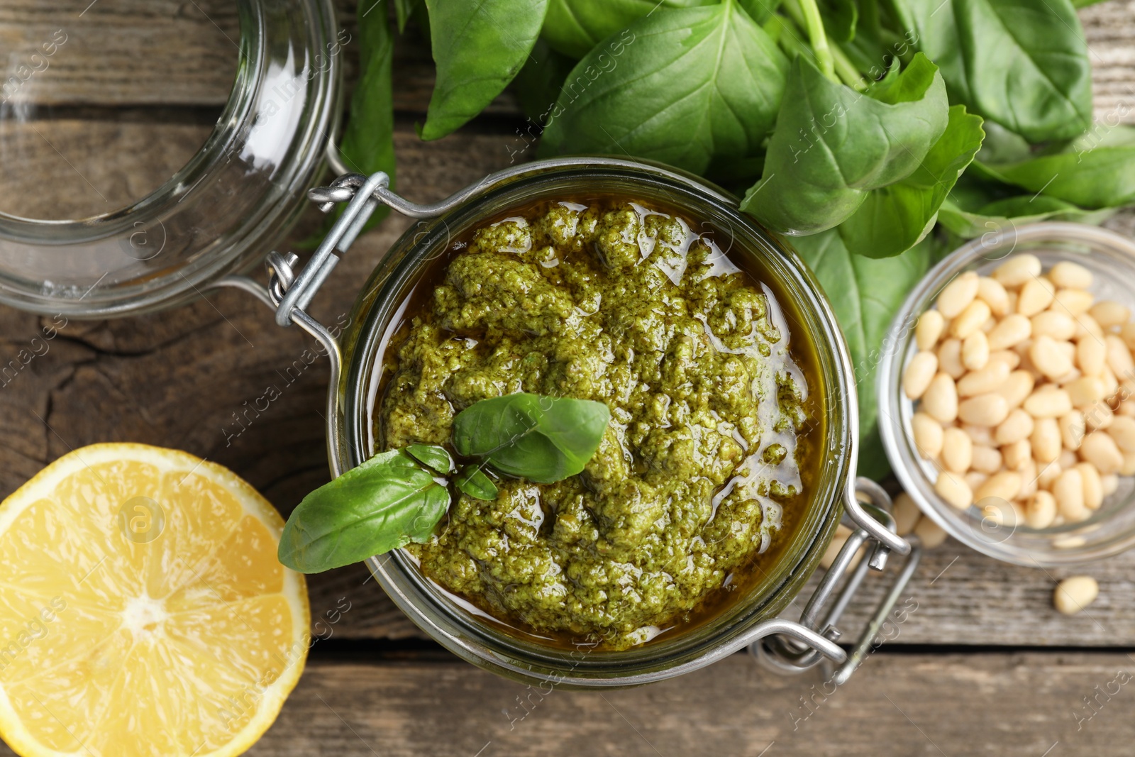 Photo of Tasty pesto sauce in jar, pine nuts, basil and lemon on wooden table, top view