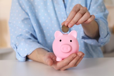 Woman putting coin into pink piggy bank at white table, closeup