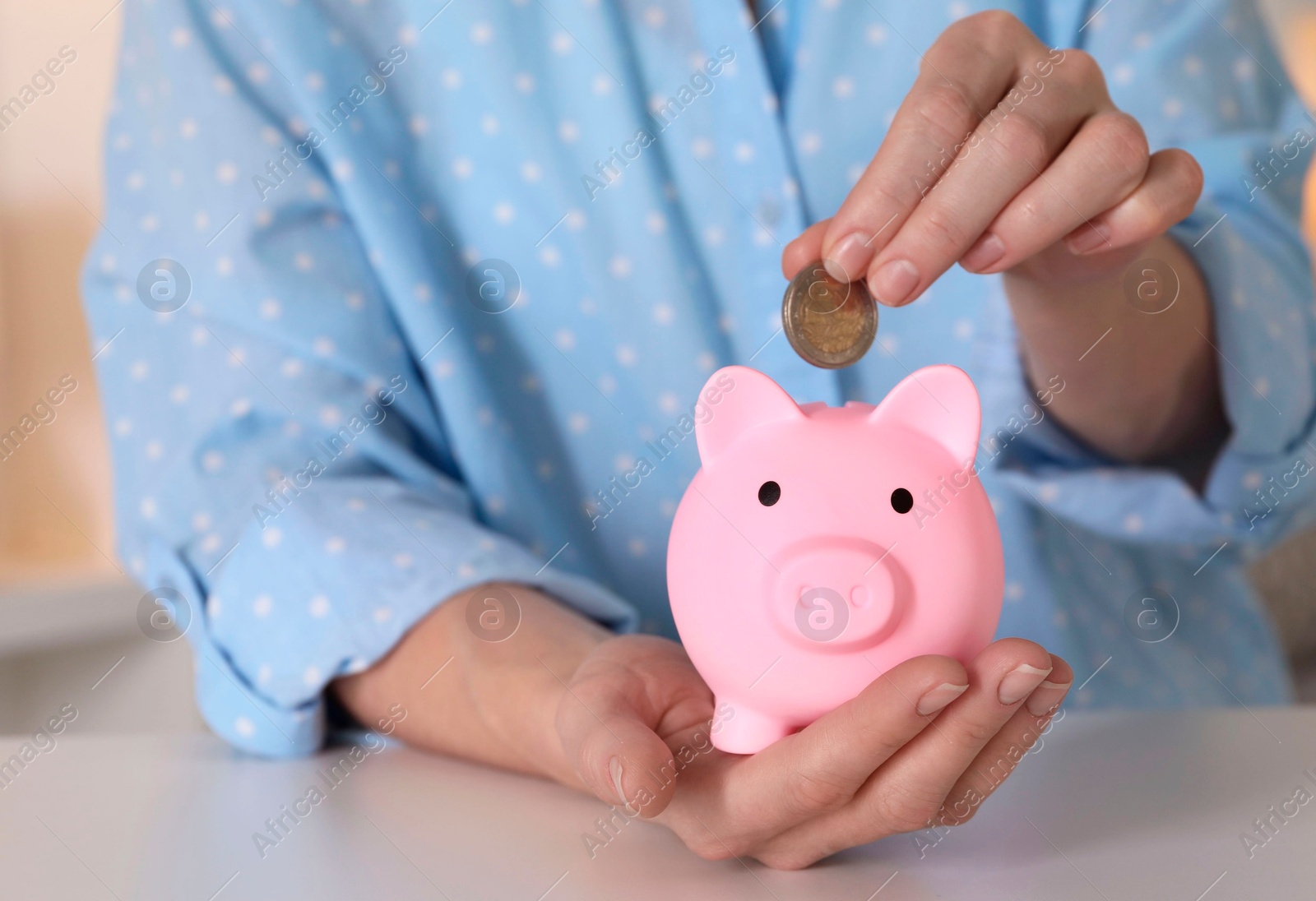 Photo of Woman putting coin into pink piggy bank at white table, closeup