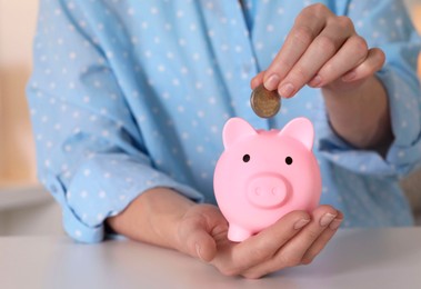 Woman putting coin into pink piggy bank at white table, closeup