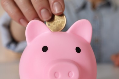 Woman putting coin into pink piggy bank at table, closeup