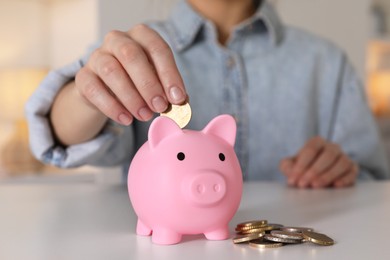 Woman putting coin into pink piggy bank at white table, closeup