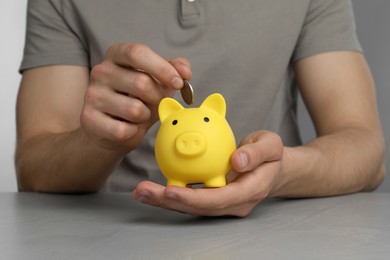 Photo of Man putting coin into yellow piggy bank at grey table, closeup