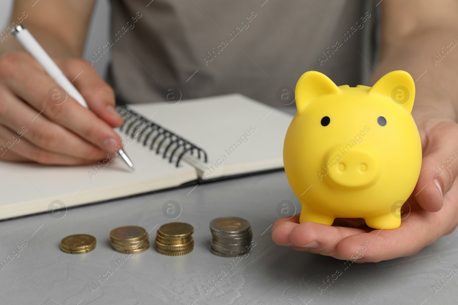 Photo of Man with yellow piggy bank, coins and notebook at grey table, closeup