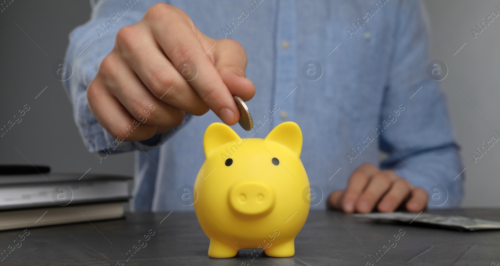 Photo of Man putting coin into yellow piggy bank at black table, closeup