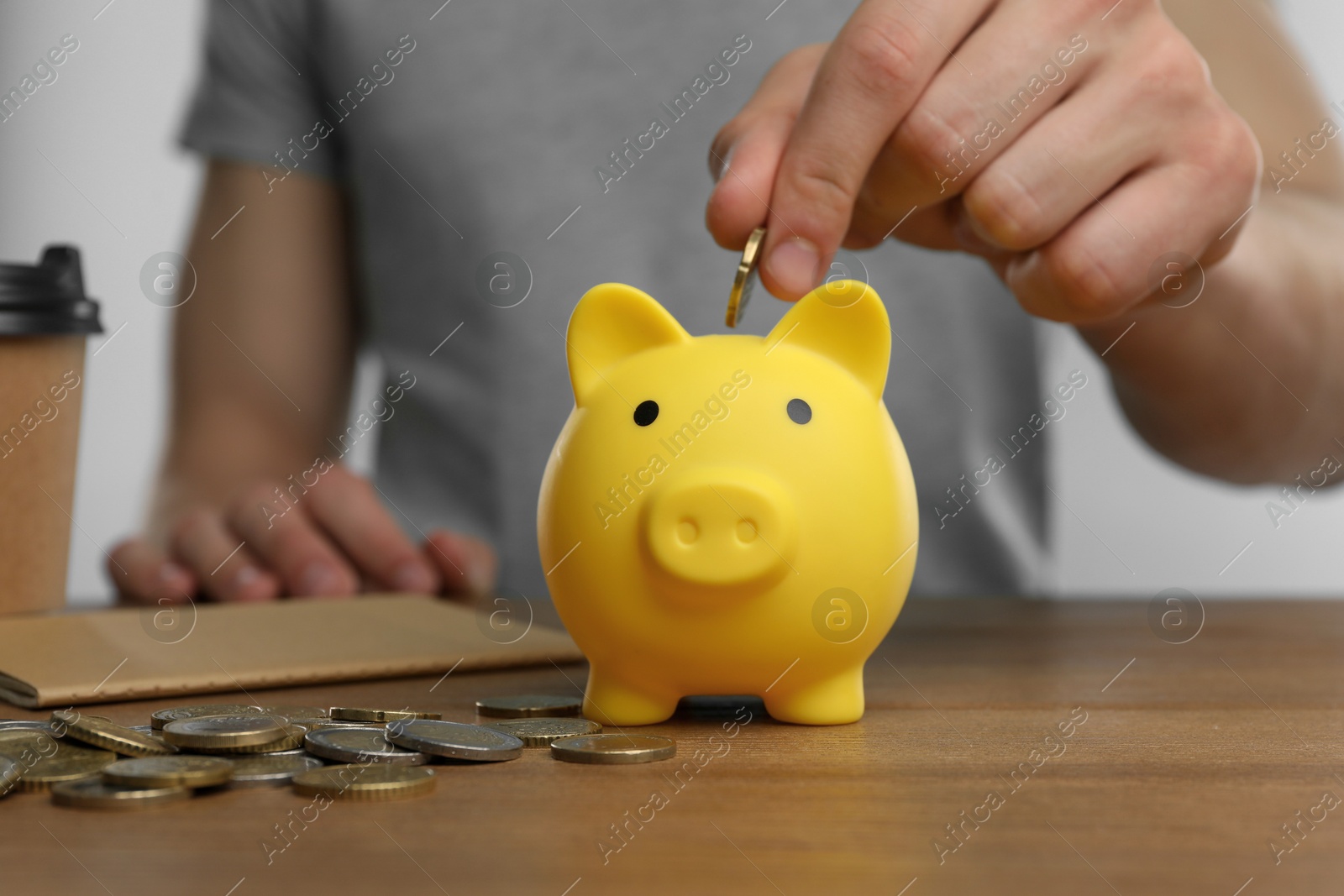 Photo of Man putting coin into yellow piggy bank at wooden table, closeup