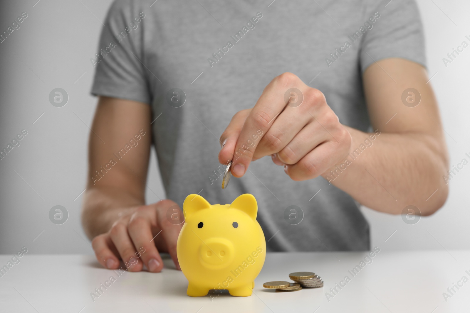 Photo of Man putting coin into yellow piggy bank at white table, closeup