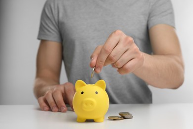 Photo of Man putting coin into yellow piggy bank at white table, closeup