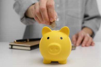 Photo of Woman putting coin into yellow piggy bank at white table, closeup