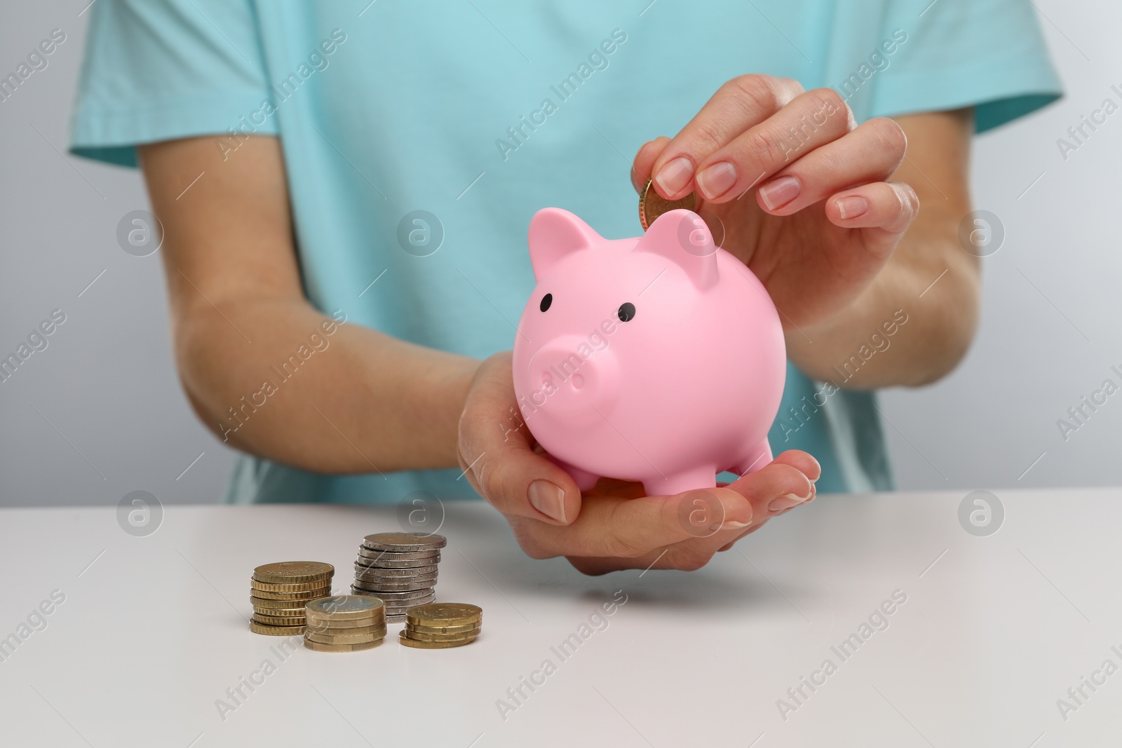 Photo of Woman putting coin into pink piggy bank at white table, closeup