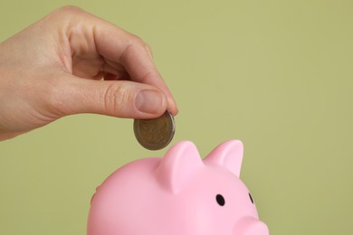 Photo of Woman putting coin into pink piggy bank on olive background, closeup. Space for text